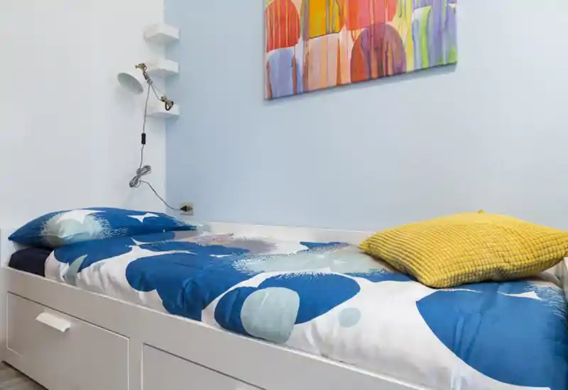 Bright single bedroom with a neatly made daybed featuring blue patterned bedding, a yellow cushion and colorful wall art — compact and welcoming sleeping space.