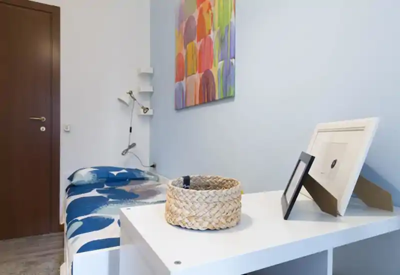 Bright, simply styled bedroom with a single bed dressed in blue patterned bedding, a bedside lamp and wall art; a white shelf/desk in the foreground holds decorative frames and a woven basket.