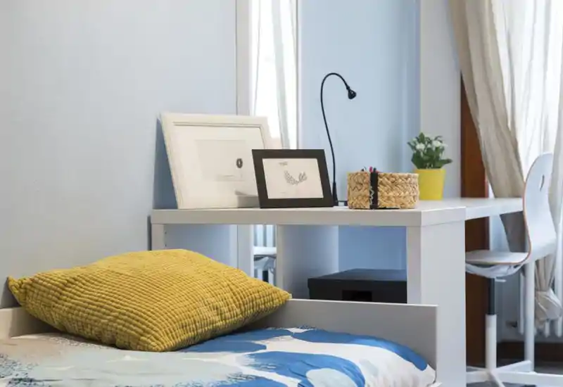 Bright, tidy bedroom corner with a visible bed, textured yellow cushion and a neat desk with framed prints — ideal for a cozy single or studio listing.