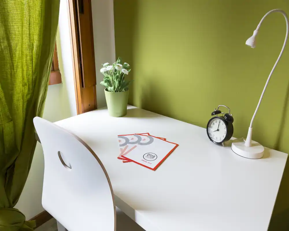 Angolo scrivania luminoso con tavolo bianco, sedia, lampada da tavolo e sveglia su parete verde; una pianta in vaso e tende verdi aggiungono un tocco accogliente.