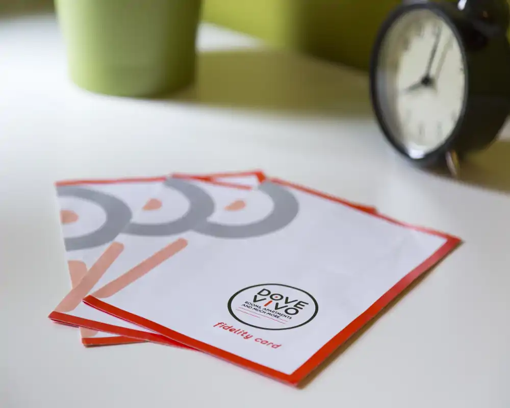 Primo piano di biglietti/fidelity card su un piano d'appoggio con una sveglia e una pianta sfocate sullo sfondo; non è visibile un contesto di stanza chiaro.