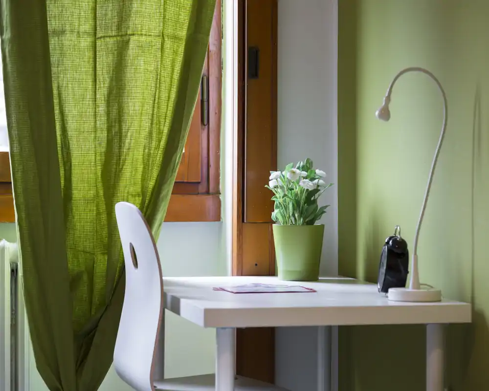 Angolo lavoro luminoso vicino alla finestra con scrivania bianca, sedia, lampada flessibile e vaso con pianta; tonalità verdi e luce naturale creano un angolo di studio tranquillo.