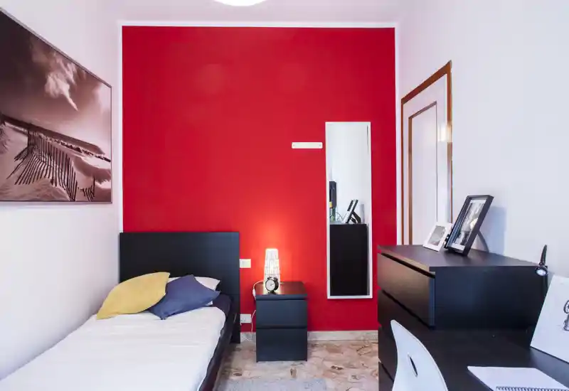 Chambre simple moderne et épurée avec un cadre de lit noir devant un mur rouge vif, table de chevet avec lampe et réveil, commode et bureau avec photos encadrées — lumineuse et bien décorée pour une annonce.