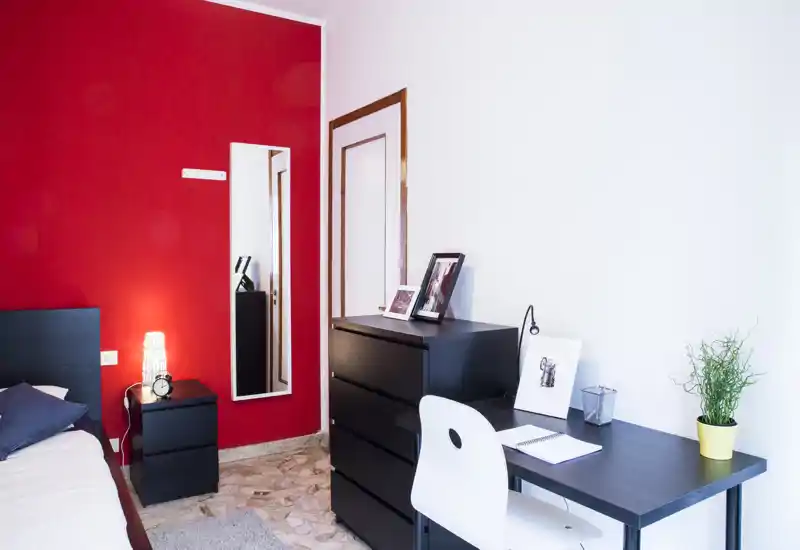 Chambre moderne et lumineuse avec lit noir et table de chevet assortie, bureau et commode contre un mur blanc et un mur d'accent rouge vif. Lampe de chevet, photos encadrées et petite plante apportent une ambiance soignée et chaleureuse.