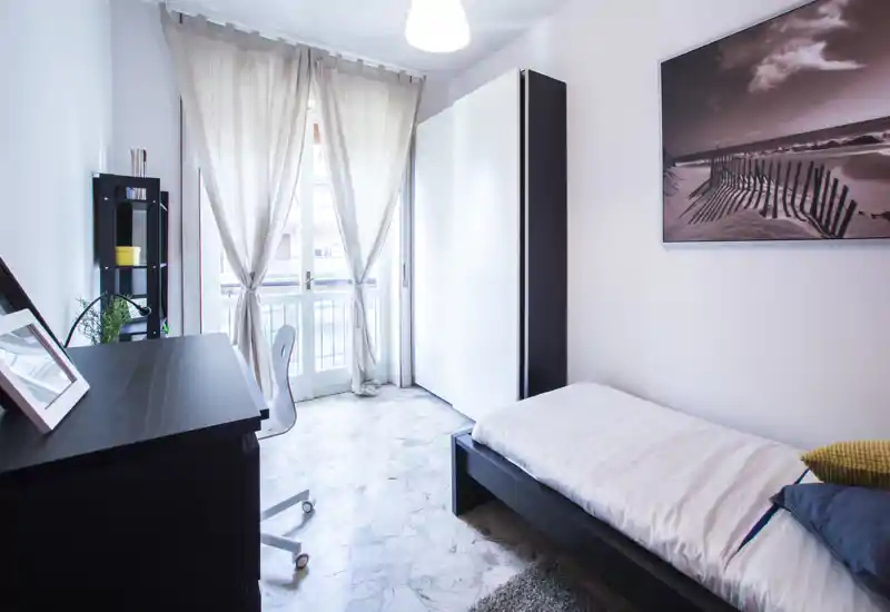 Chambre simple moderne et lumineuse avec lit bas à cadre noir, linge de lit blanc, bureau près de la porte du balcon et décoration minimaliste — idéale pour étudiant ou professionnel.