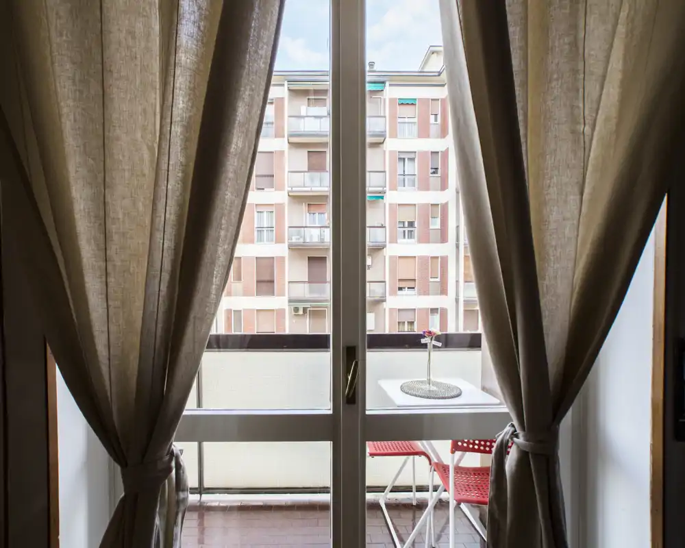 Petit balcon privé avec une table blanche compacte et deux chaises rouges, vu à travers des rideaux beiges attachés. Idéal pour un café du matin avec vue urbaine.