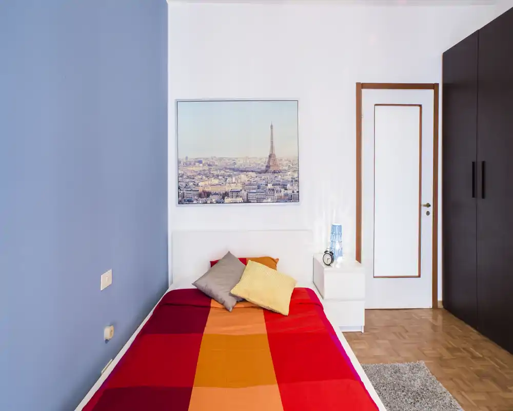 Chambre simple lumineuse et soignée avec une couette colorée, une table de chevet et une armoire ; un cadre représentant la ville au-dessus du lit apporte du caractère.