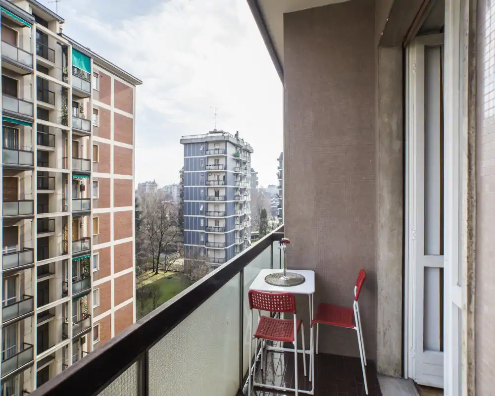 Balcon étroit et ensoleillé avec petite table et deux chaises rouges, donnant sur des immeubles voisins et un parc — idéal pour un café matinal.