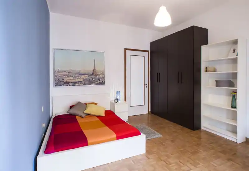 Chambre lumineuse et bien rangée avec un lit coloré, une table de chevet et une grande armoire foncée. Sol en parquet, cadre mural et étagère simple — idéale pour un séjour privé et confortable.