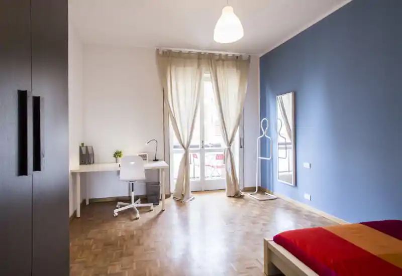 Chambre lumineuse et moderne avec bureau et accès au balcon. Sol en parquet, mur bleu en accent, armoire simple et lit coloré apportant une touche accueillante.