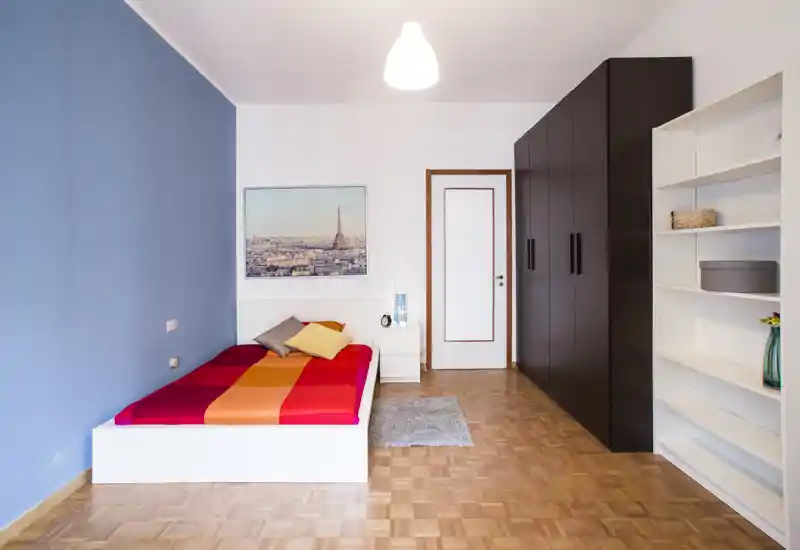Chambre simple lumineuse et moderne avec un lit plateforme blanc aux couettes colorées, une table de chevet avec lampe, une grande armoire foncée et des étagères ouvertes; parquet soigné et une affiche encadrée pour un espace bien stylé.