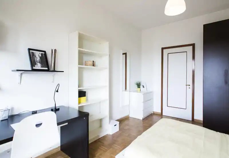 Bright, minimal bedroom with a partial view of the bed, a desk with shelving and wardrobe — tidy modern styling ideal for a single occupant.