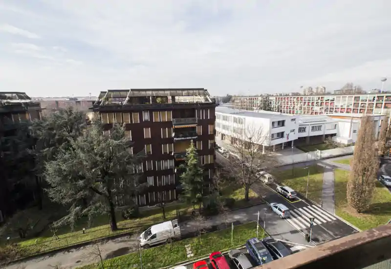 Urban balcony view showing neighboring apartment blocks, street, parked cars and green trees — bright, wide-angle exterior shot ideal to showcase the view from the unit.