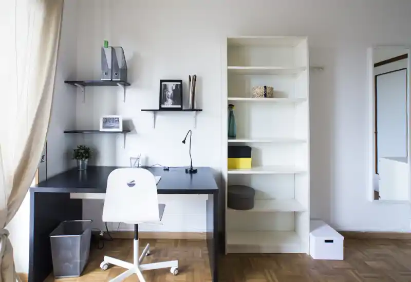 Bright, minimalist home office corner with a black desk, white chair and shelving; tidy workspace with framed photos, a small plant and a tall white bookcase.
