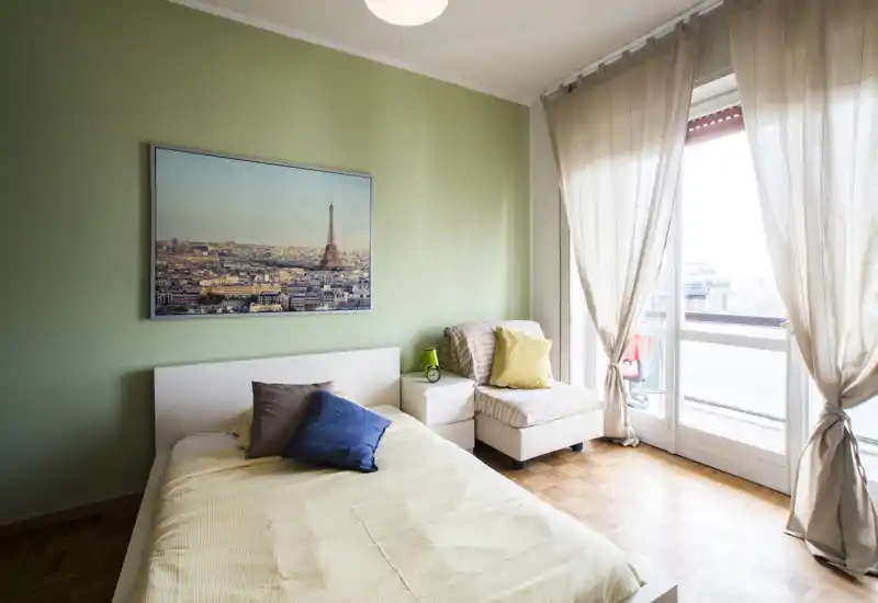 Chambre lumineuse et bien agencée avec un lit double, coussins moelleux et un coin siège près d'une grande fenêtre donnant sur un balcon. Mur vert pâle et lumière naturelle pour une ambiance calme et chaleureuse.
