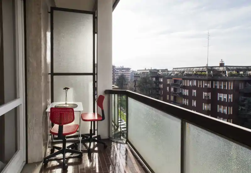 Sunny narrow balcony with a small table and two red chairs, glossy tiled floor and a city view — a cozy spot for morning coffee.