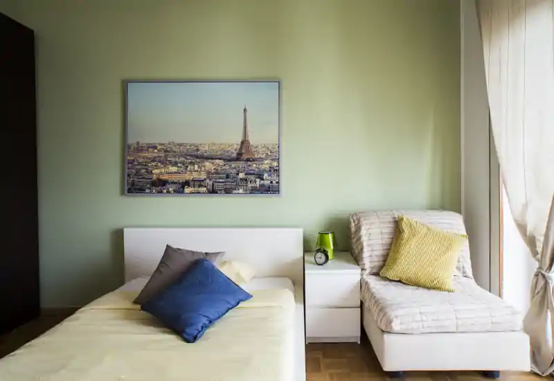 Chambre lumineuse et soignée avec un lit simple, une table de chevet et un confortable jourbed près d'une fenêtre ensoleillée ; murs verts pâles et tableau encadré pour une atmosphère apaisante.