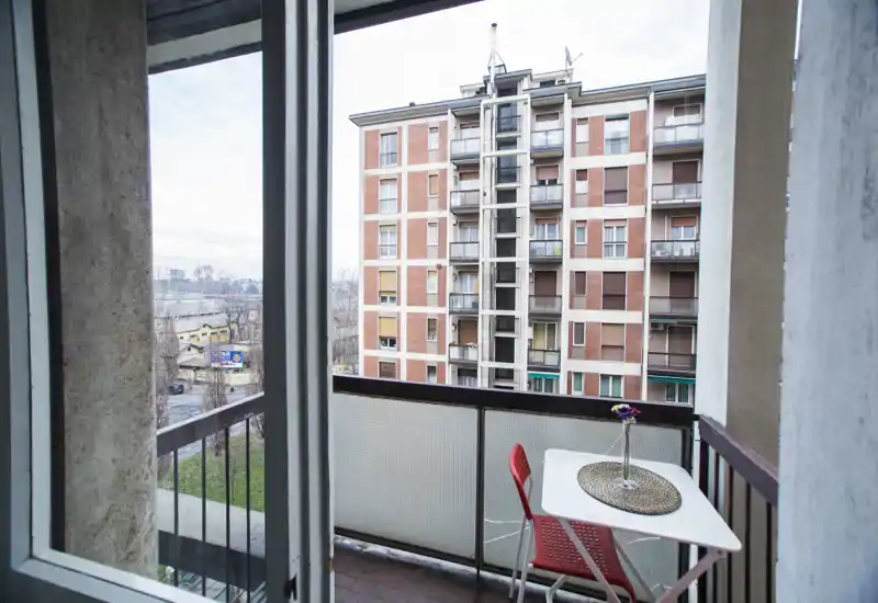 Small furnished balcony with a table, red chair and railing; view onto neighboring apartment building and urban greenery. Bright natural light and simple outdoor seating ideal for morning coffee.