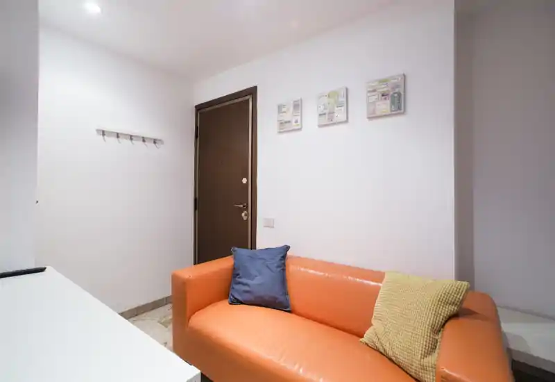 Small, well-lit entrance with a bright orange two-seater sofa, decorative cushions and a wooden entry door — a welcoming foyer area with clean, modern styling.