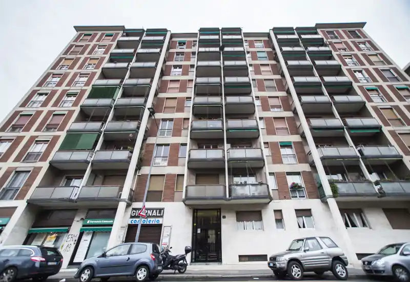Street-level view of a multi-story residential building with balconies, ground-floor shops and parked cars — ideal for showing the building exterior and neighborhood context.