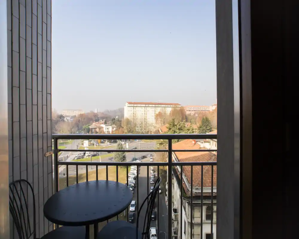 Balcone soleggiato con piccolo tavolo rotondo e due sedie in metallo, affacciato su una strada alberata e palazzi residenziali in lontananza — ideale per il caffè mattutino.