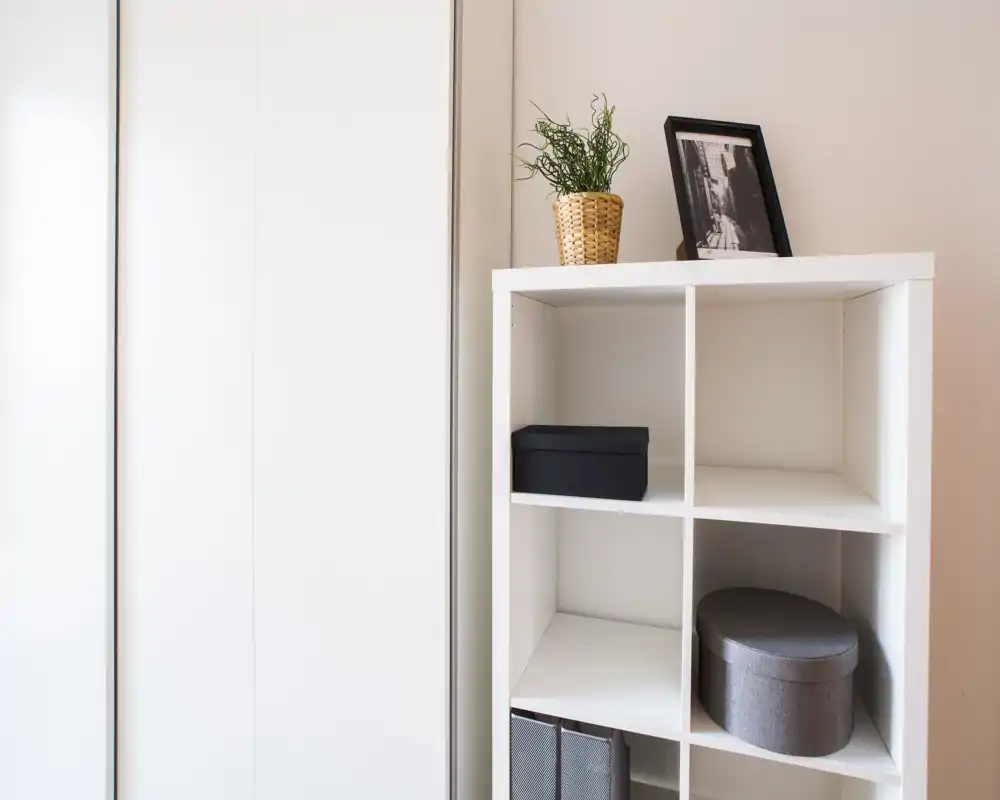 Primo piano di un mobile a cubi bianco accanto a una porta armadio scorrevole; scatola decorativa, contenitore rotondo e una piccola pianta sopra. La foto mostra l'arredo contenitore più che l'ambiente completo.