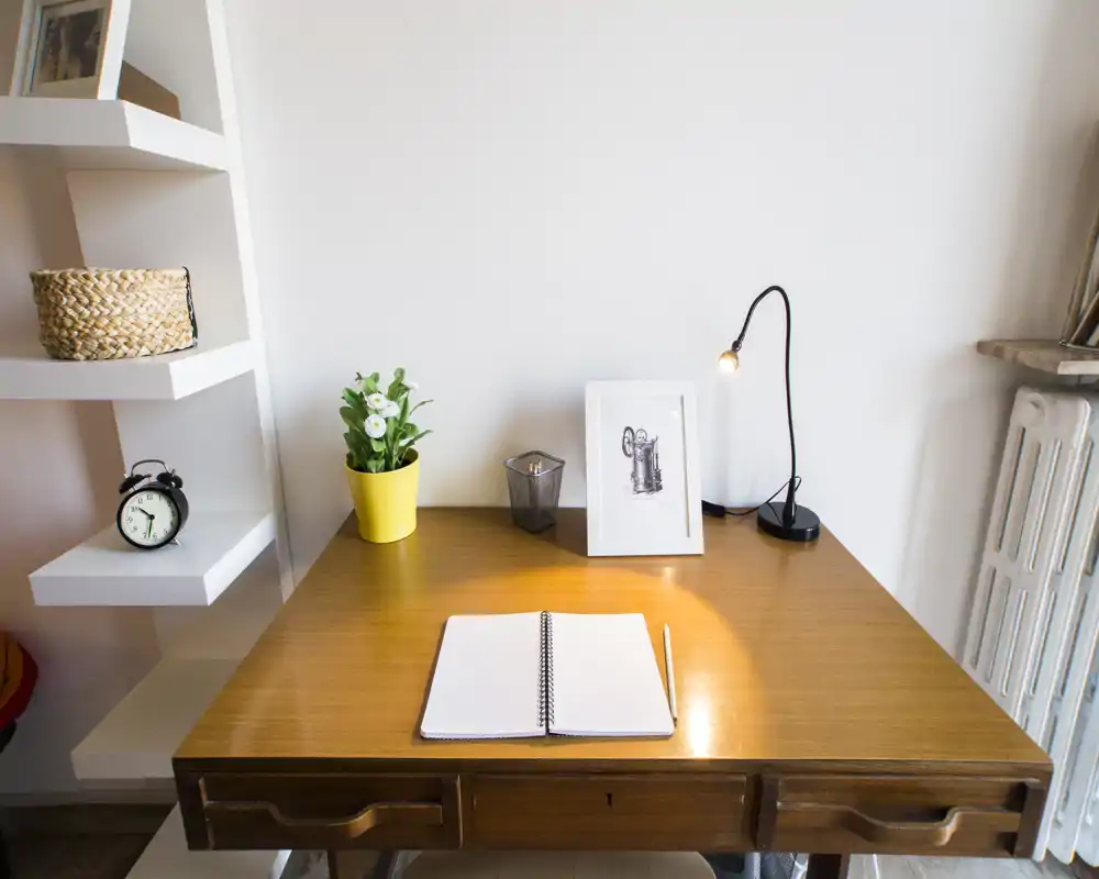 Angolo scrivania luminoso e ordinato con piano in legno, lampada da tavolo, piccola pianta e quadro incorniciato — perfetto per lavorare o leggere.