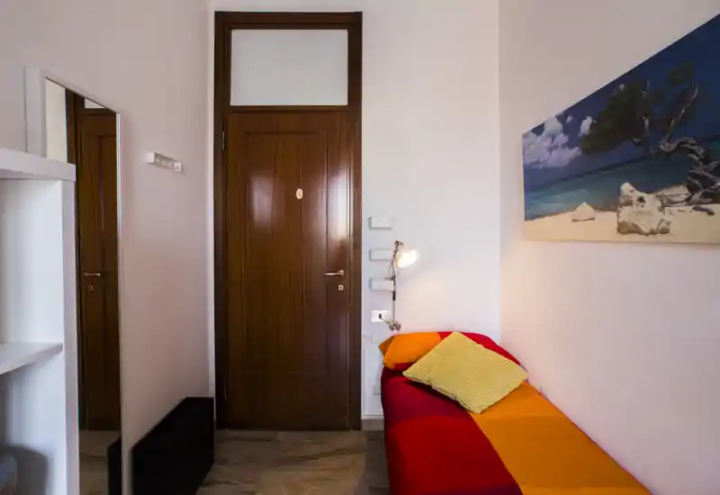 Camera da letto compatta con un letto singolo colorato, cuscini morbidi e una lampada da lettura accanto alla testiera. Un quadro e uno specchio a figura intera aggiungono profondità all'ambiente.