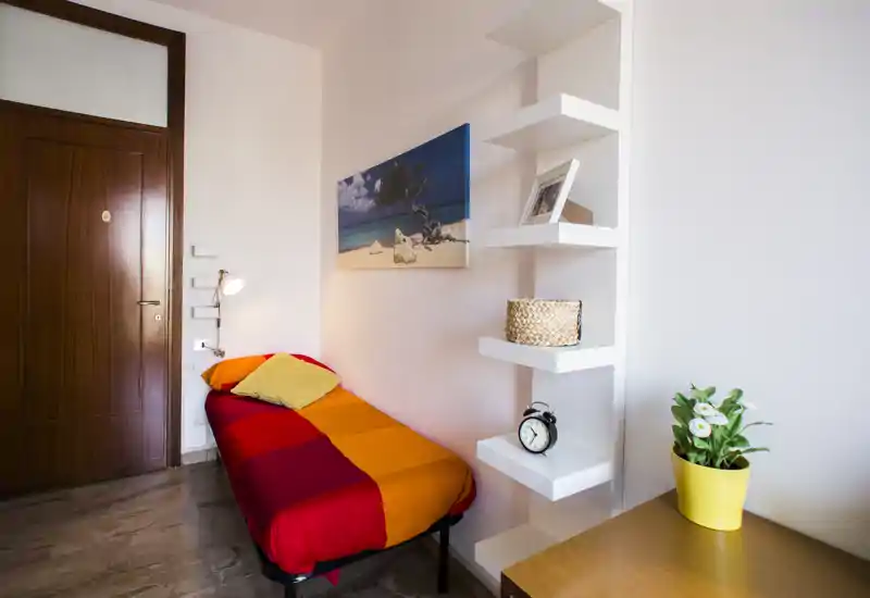 Camera singola compatta con letto singolo dai colori vivaci arancione e rosso, lampada da lettura, mensole a sbalzo e una piccola scrivania con pianta. Spazio luminoso e ben allestito, ideale per un affitto per una persona o breve periodo.