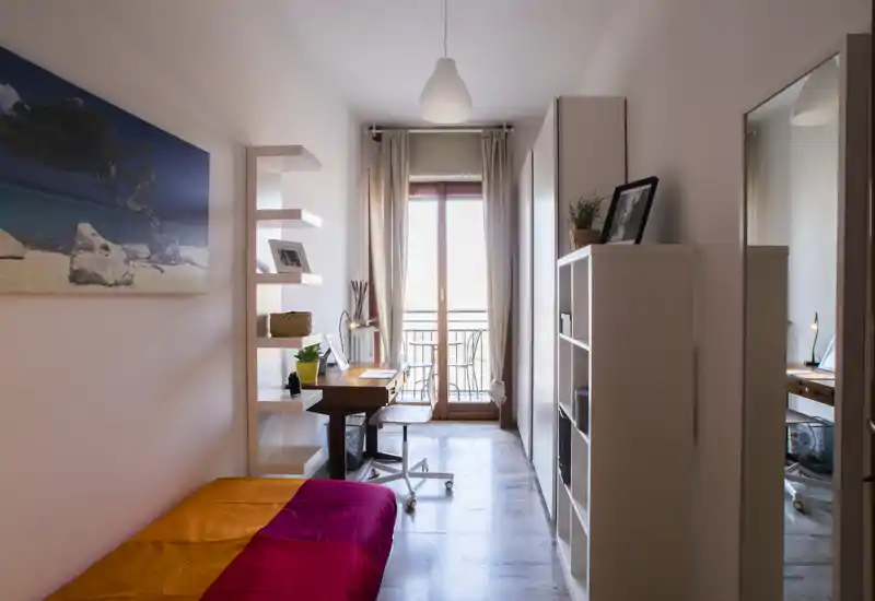 Camera compatta con letto singolo colorato, scaffalature e una scrivania davanti a una porta finestra luminosa — ideale per uno studente o una persona sola che cerca uno spazio ordinato e ben illuminato.