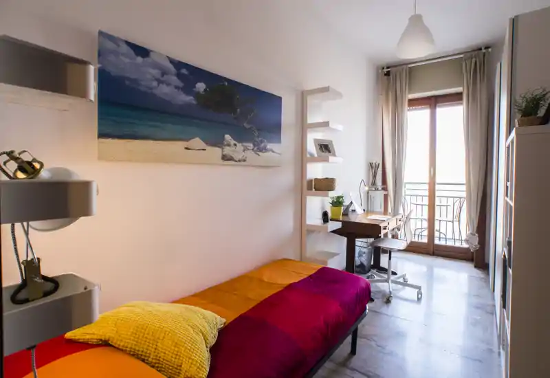Camera da letto singola, luminosa e accogliente con biancheria colorata, scrivania vicino alla porta-balcone e quadro decorativo. La luce naturale entra dalla finestra a tutta altezza, creando un ambiente caldo e adatto a studenti o single.