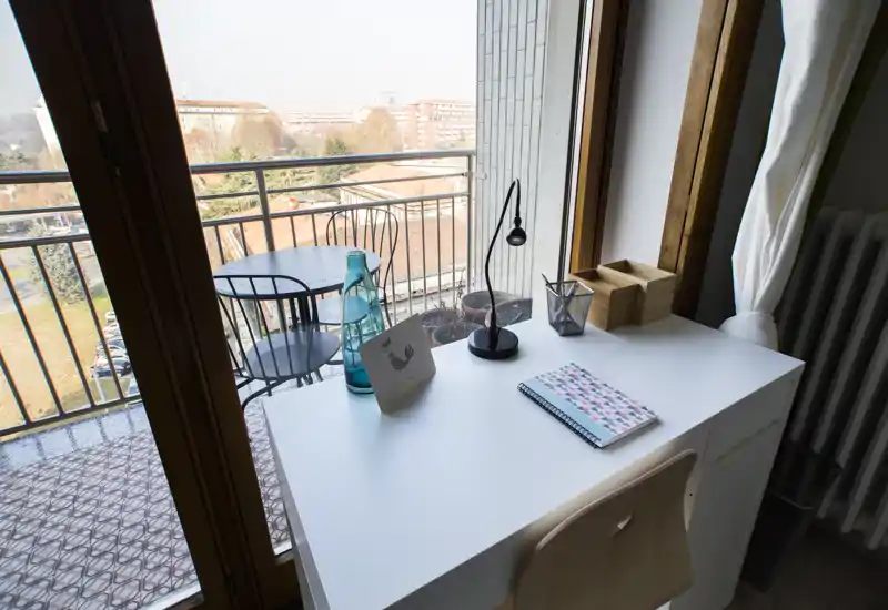 Piccolo balcone con scrivania bianca vicino alla porta finestra, un tavolino bistro con sedie e alcuni vasi: vista cittadina luminosa perfetta per colazione o pause di lavoro.