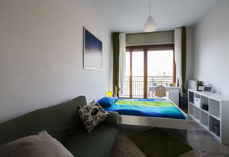 Camera da letto luminosa e semplice con un letto matrimoniale vicino alla porta finestra che dà sul balcone. La stanza presenta una scrivania, una libreria e un divano confortevole, con tessuti verdi e blu che creano un’atmosfera fresca e accogliente.