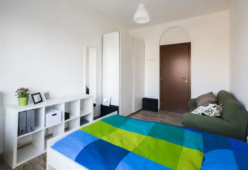 Camera da letto luminosa e moderna con copriletto blu-verde, scaffalatura compatta, armadi bianchi alti e un piccolo divano verde—stile pulito e ottima luce naturale.