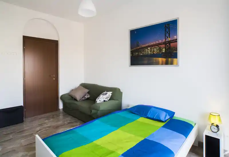Camera da letto luminosa e semplice con un letto singolo colorato, una lampada da comodino e un piccolo divanetto verde. Linee pulite, quadro con paesaggio urbano e pareti neutre per un'atmosfera moderna e tranquilla.