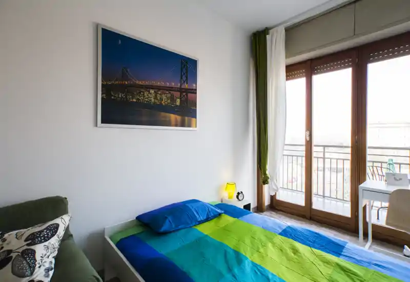 Camera da letto luminosa e ben arredata con letto matrimoniale in lenzuola blu e verdi, lampada da comodino e grandi porte finestra che rendono la stanza molto luminosa.