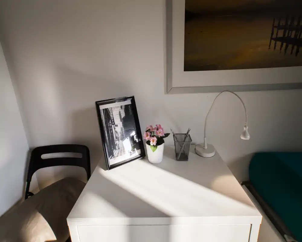 Compact bedroom corner with a bedside table featuring a lamp, framed photo and small plant; partial view of the mattress at the right edge and a chair to the left, lit by warm natural light.
