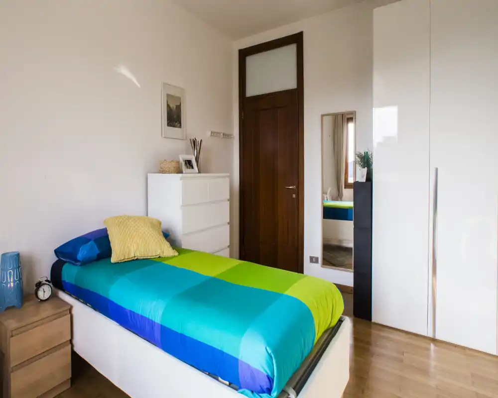 Bright, neatly staged single bedroom with a colorful blue-green duvet, bedside table and wardrobe. The room features natural light, clean wooden floors and minimal modern furnishings suitable for a rental listing.