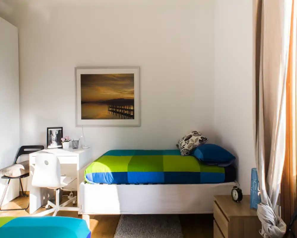 Sunny, neatly styled single bedroom with a colorful bedspread, bedside table, desk and natural light from a window. The room feels bright and welcoming — ideal for a rental listing highlight.