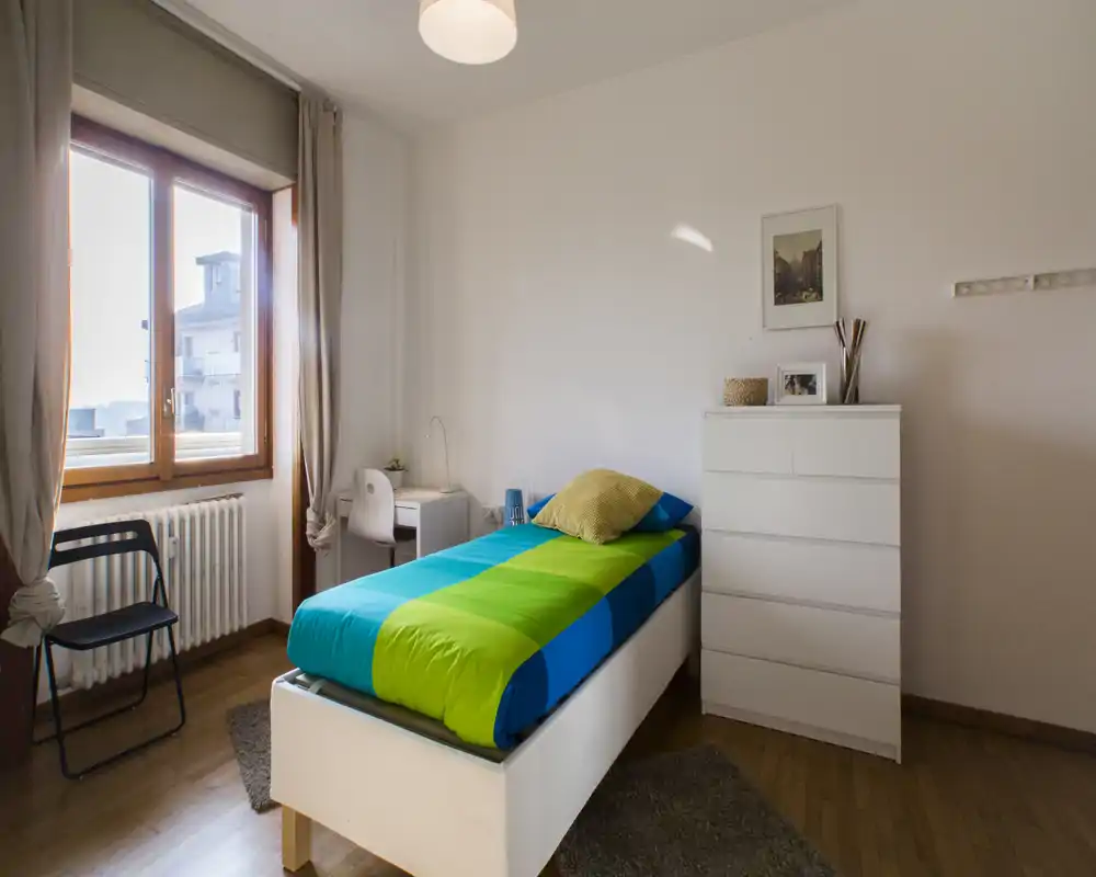 Sunny single bedroom with a colorful blue-and-green bed, white chest of drawers and a small desk by the window; wooden floors and soft natural light create a welcoming atmosphere.