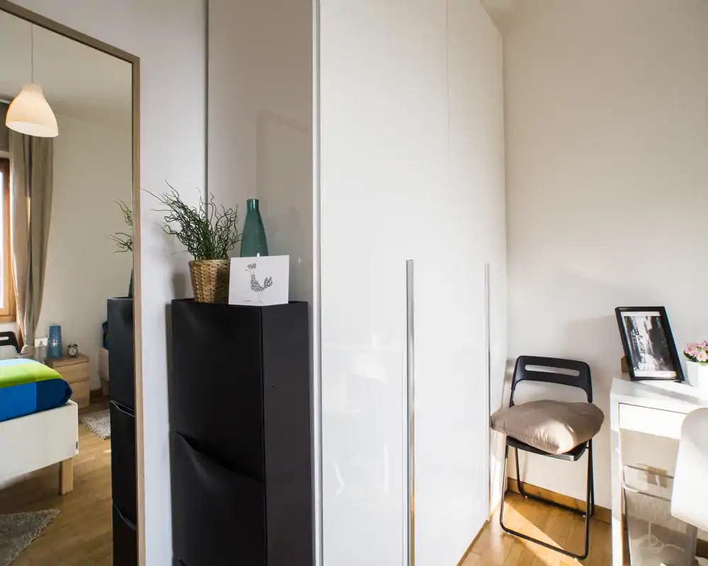 Bright, modern bedroom corner with a visible bed reflected in a large mirror, a glossy white wardrobe, and a small desk area with a chair — warm wooden floors and tasteful decorative accents.