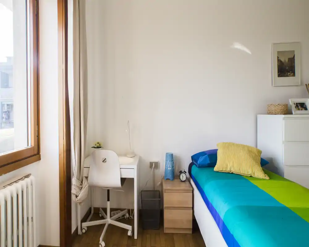 Bright single bedroom with a colorful turquoise and green bed, bedside table with lamp and clock, small study desk by the window and soft natural light coming through large windows.