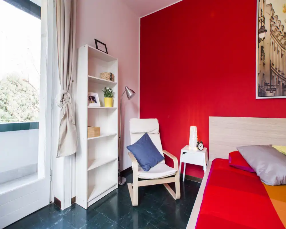 Camera da letto moderna e luminosa con parete rossa, letto parzialmente visibile, poltrona accogliente e piccolo comodino vicino alla porta del balcone che lascia entrare luce naturale.