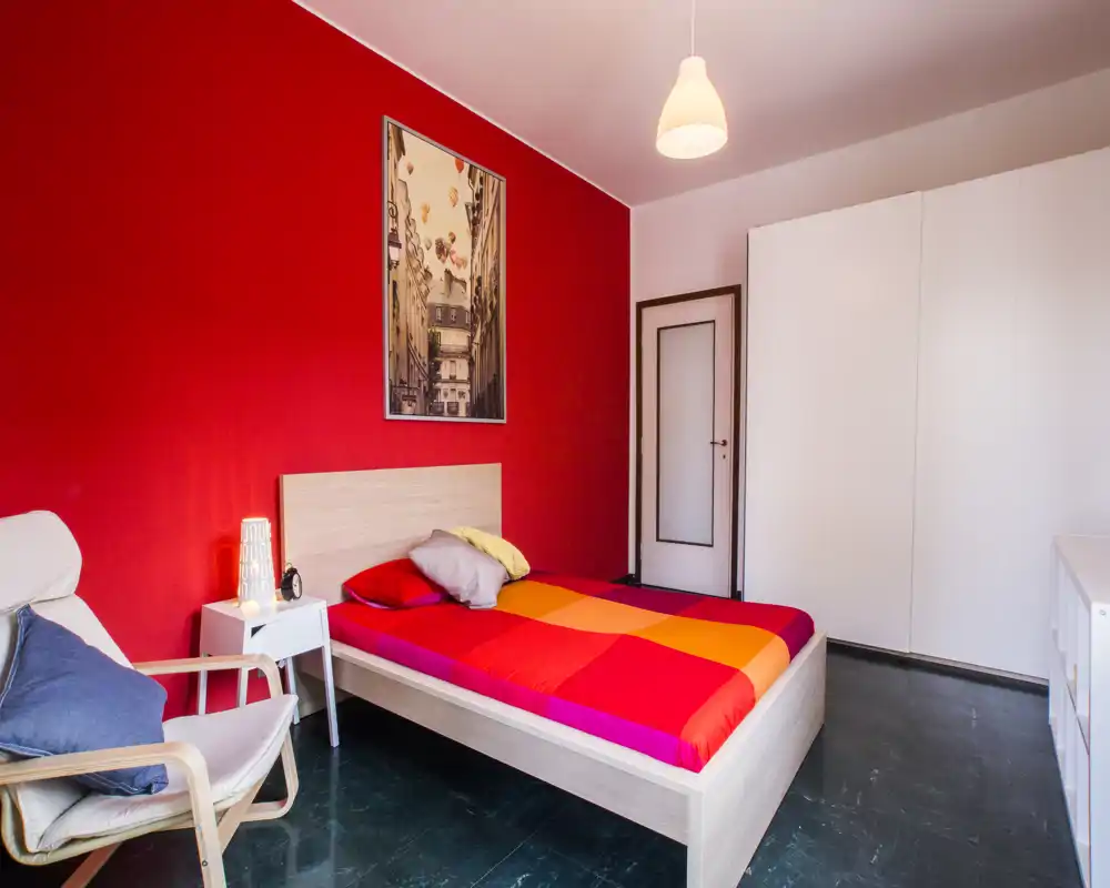 Camera da letto moderna e luminosa con un letto colorato contro una parete rossa, comodino con lampada e una poltrona confortevole.