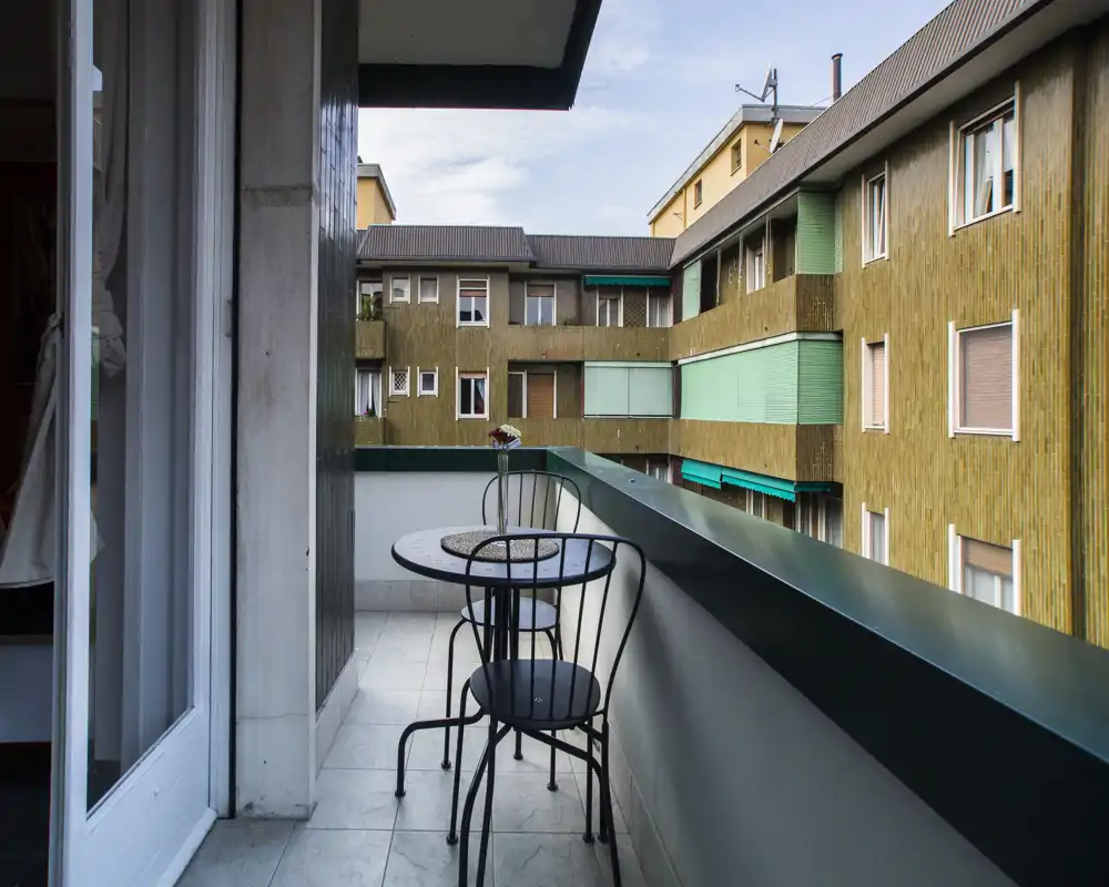 Piccolo balcone soleggiato con un tavolino bistro rotondo e due sedie in metallo, pavimento piastrellato e vista sugli edifici vicini — ideale per il caffè del mattino.