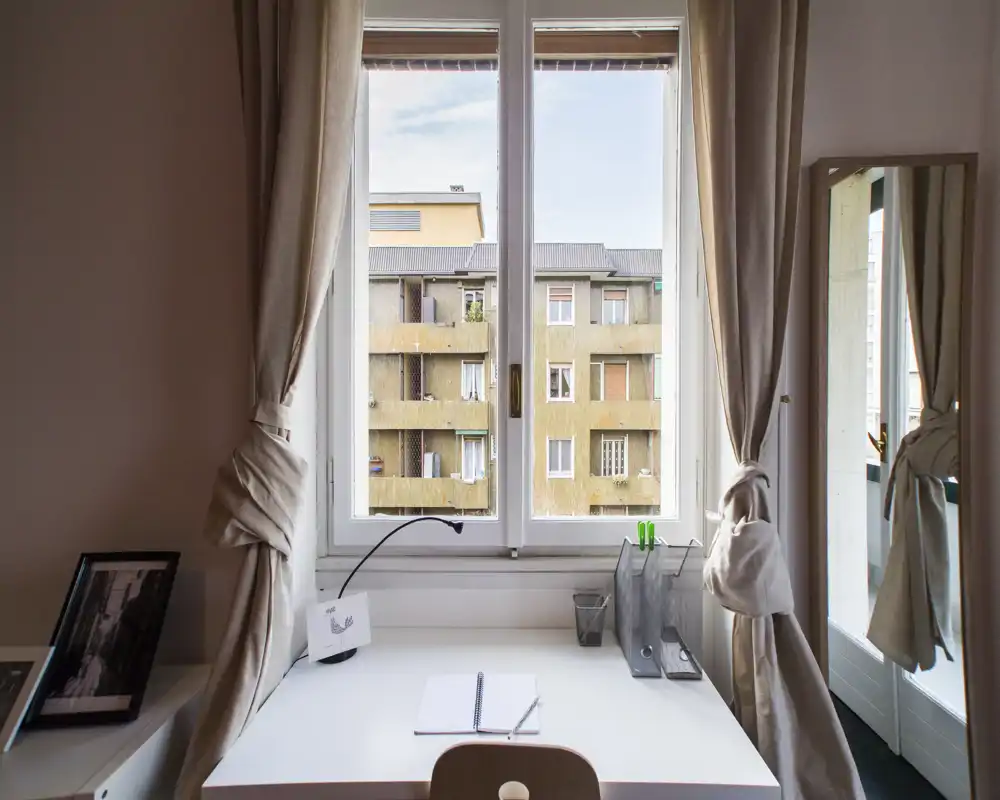 Stanza luminosa con una scrivania ordinata sotto una finestra ampia; tende neutre, uno specchio e semplici accessori da scrivania creano un angolo di lavoro tranquillo e ben illuminato con vista urbana.