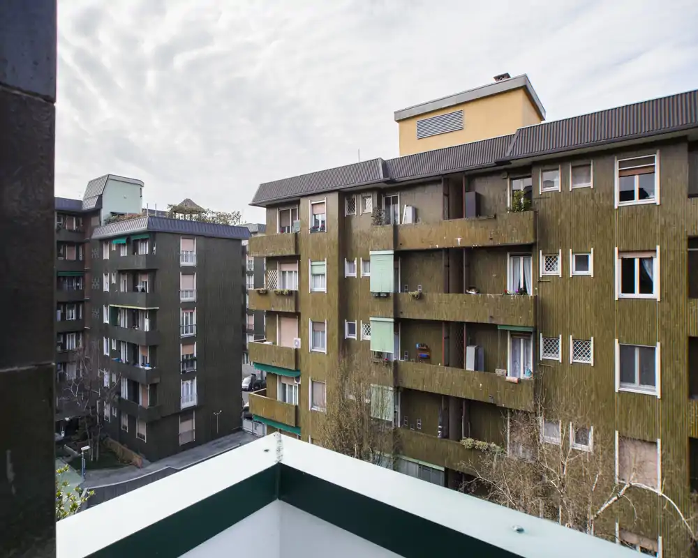 Vista dal balcone verso gli edifici condominiali circostanti, con una piccola porzione del parapetto in primo piano; cielo coperto e contesto urbano.