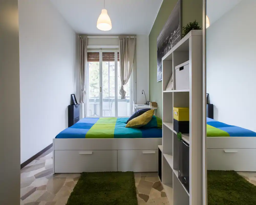 Camera da letto moderna e luminosa con letto matrimoniale ben rifatto, biancheria colorata e porta-finestra verso il balcone che illumina l’ambiente. Cassetti contenitori sotto il letto e una libreria con scatole decorative rendono lo spazio ordinato e accogliente.