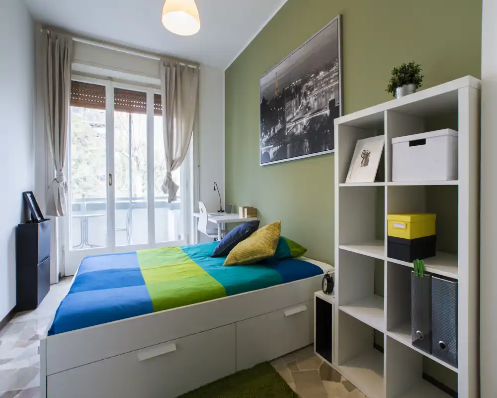 Camera da letto luminosa e moderna con letto matrimoniale, biancheria colorata, libreria a cubi e scrivania vicino alla grande finestra con balcone. Parete accent color verde e abbondante luce naturale.