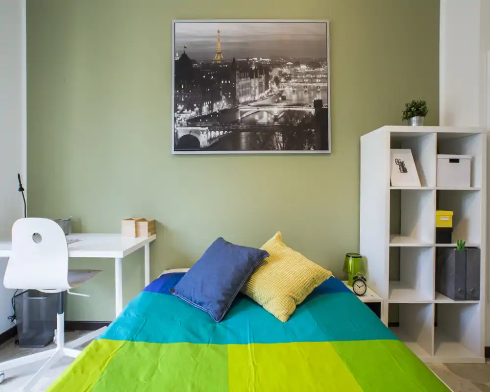 Camera da letto moderna e luminosa con letto doppio coperto da biancheria colorata, cuscini decorativi, scrivania a sinistra e libreria a cubi a destra — ordinata e ben illuminata.